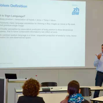 Hörsaal. Andri Reichenbacher präsentiert seinen Vortrag. Im Hintergrund eine seiner Vortragsfolien. Sie trägt den Titel: Problem Definition. What is Sign Language?