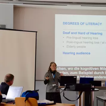 Hörsaal. Daniela Eichmeyer präsentiert ihren Vortrag. Im Hintergrund eine ihrer Vortragsfolien. Sie trägt den Titel: Degrees of Literacy. Deaf and Hard of Hearing.