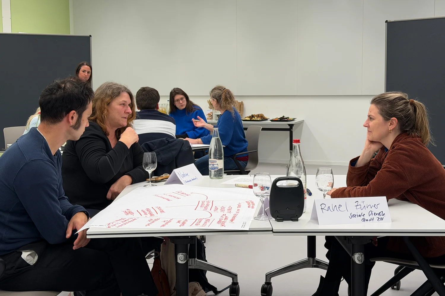 Im Unterrichtsraum sind Teilnehmende eines Workshops zu sehen, die an ihren Tischen sitzen und miteinander angeregt diskutieren.