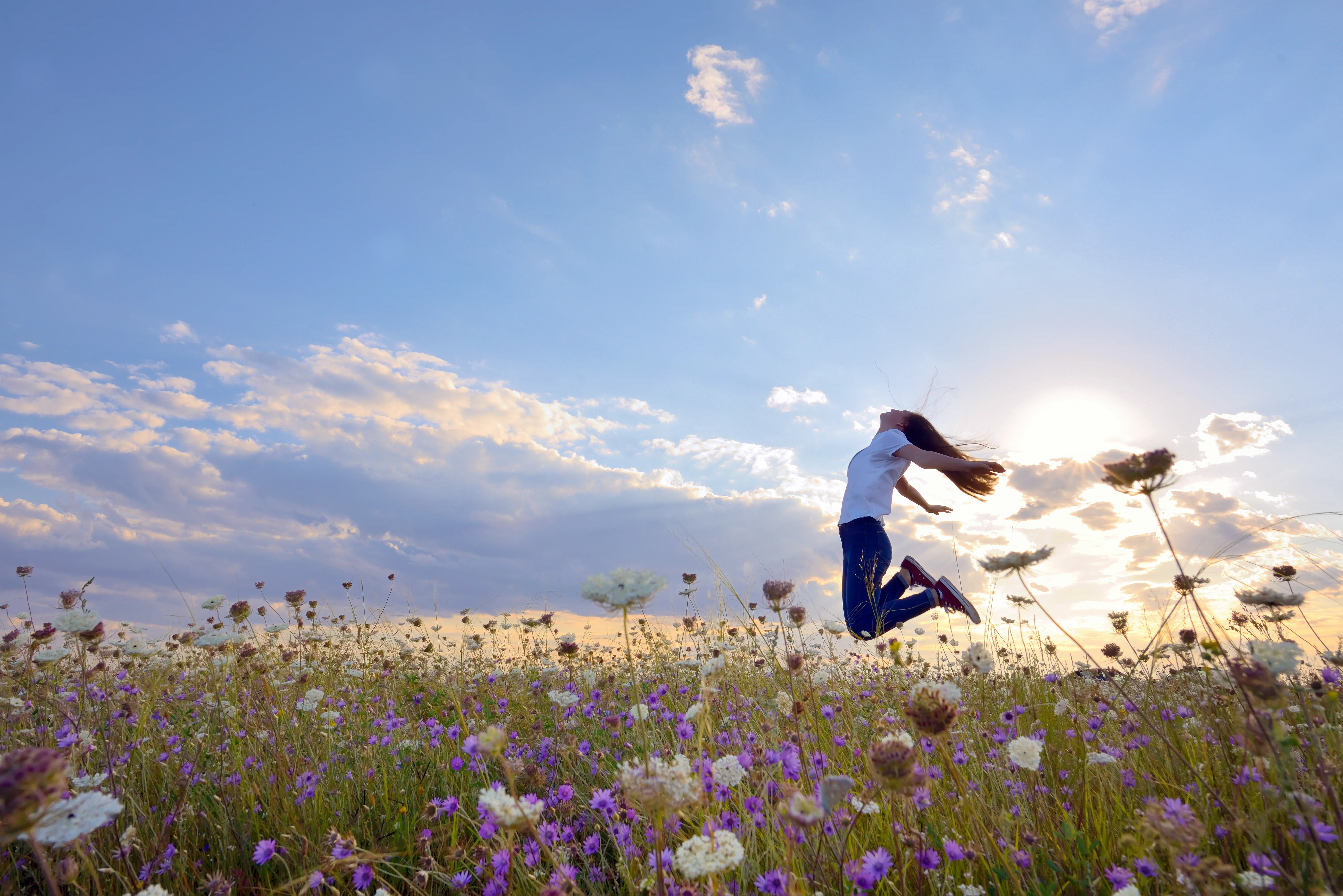 Eine junge Frau springt mit ausgebreiteten Armen über eine blühende Blumenwiese, im Hintergrund ein weiter Himmel mit Wolken.