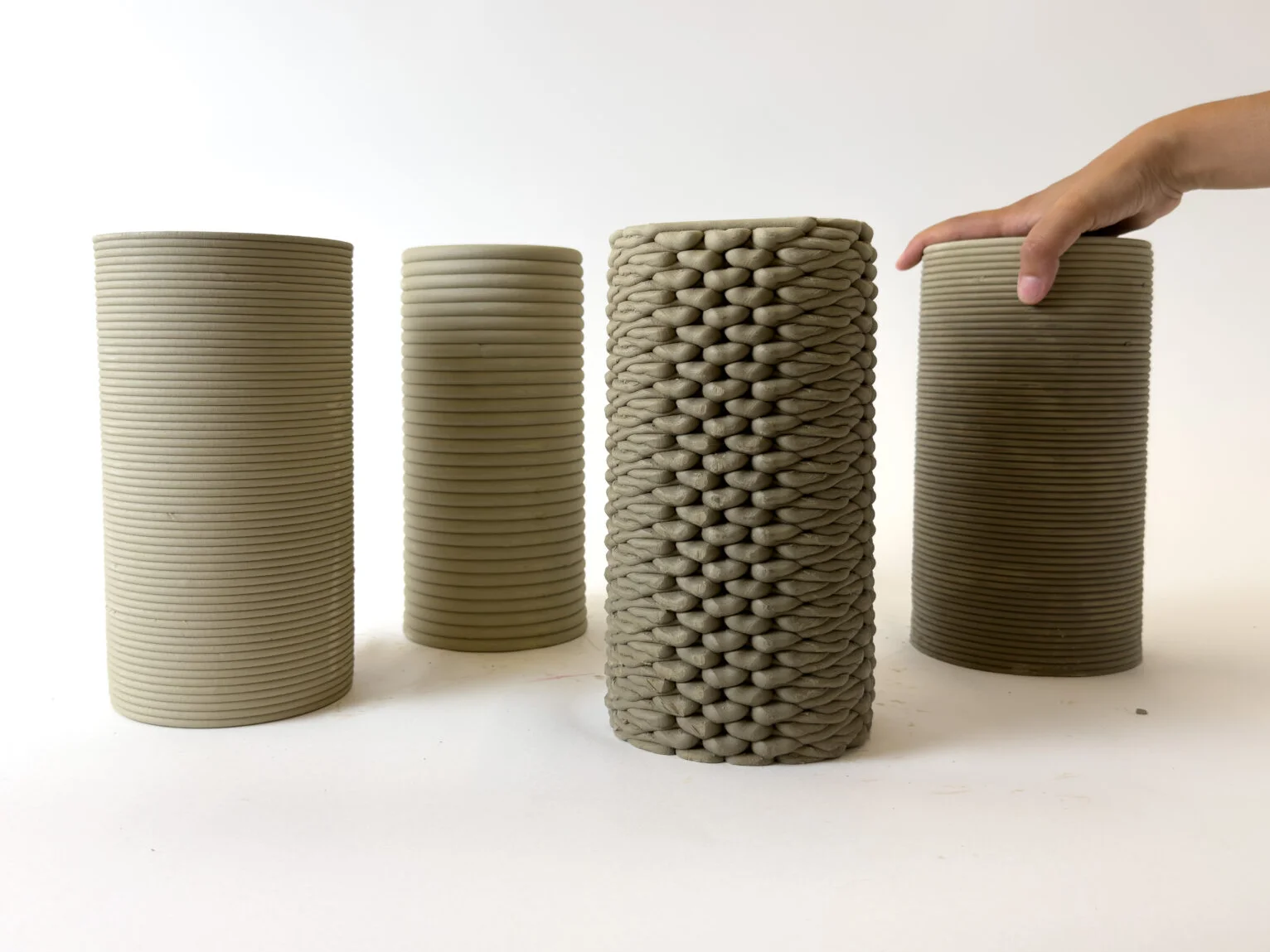 Vier 3D-gedruckte Lehmzylinder mit verschiedenen Oberflächen, einer mit Geflechtmuster; eine Hand liegt auf einem