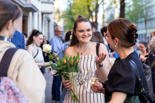 Bild vergrössern: Eine Absolventin steht vor dem Volkshaus mit anderen Personen zusammen und hält einen Blumenstrauss.