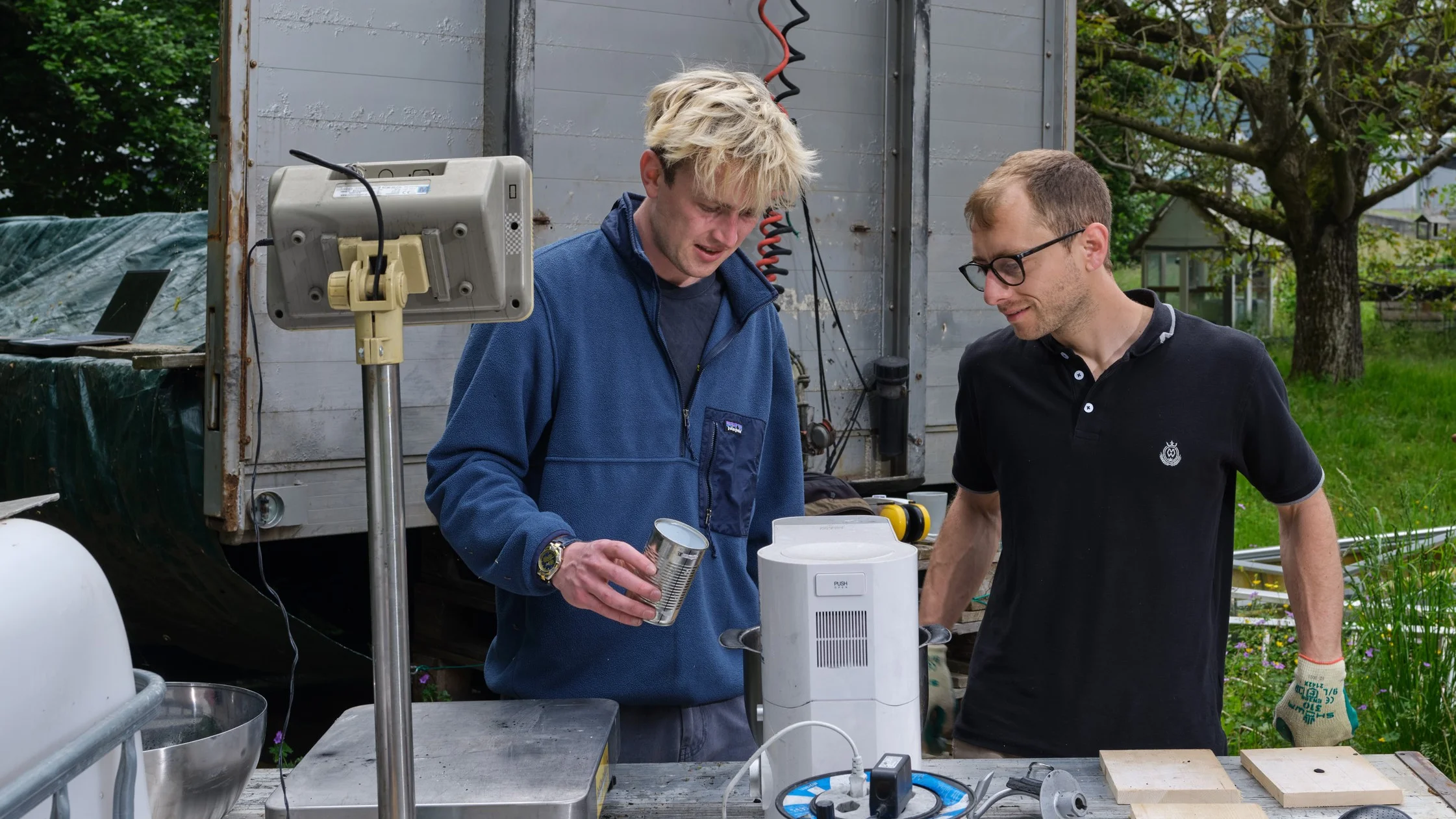 Two people stand at an outdoor work table with measuring equipment and containers, one holding a cup of material, with a truck and green outdoor space in the background.