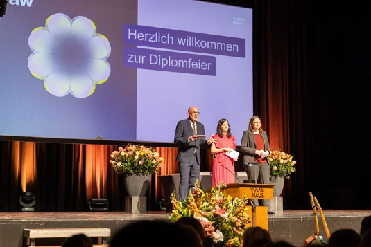 Bild vergrössern: Die drei Studiengangleitungen stehen auf der mit Blumen dekorierten Bühne. Hinter ihnen ist das grosses Wilkommens-Slide zu sehen.