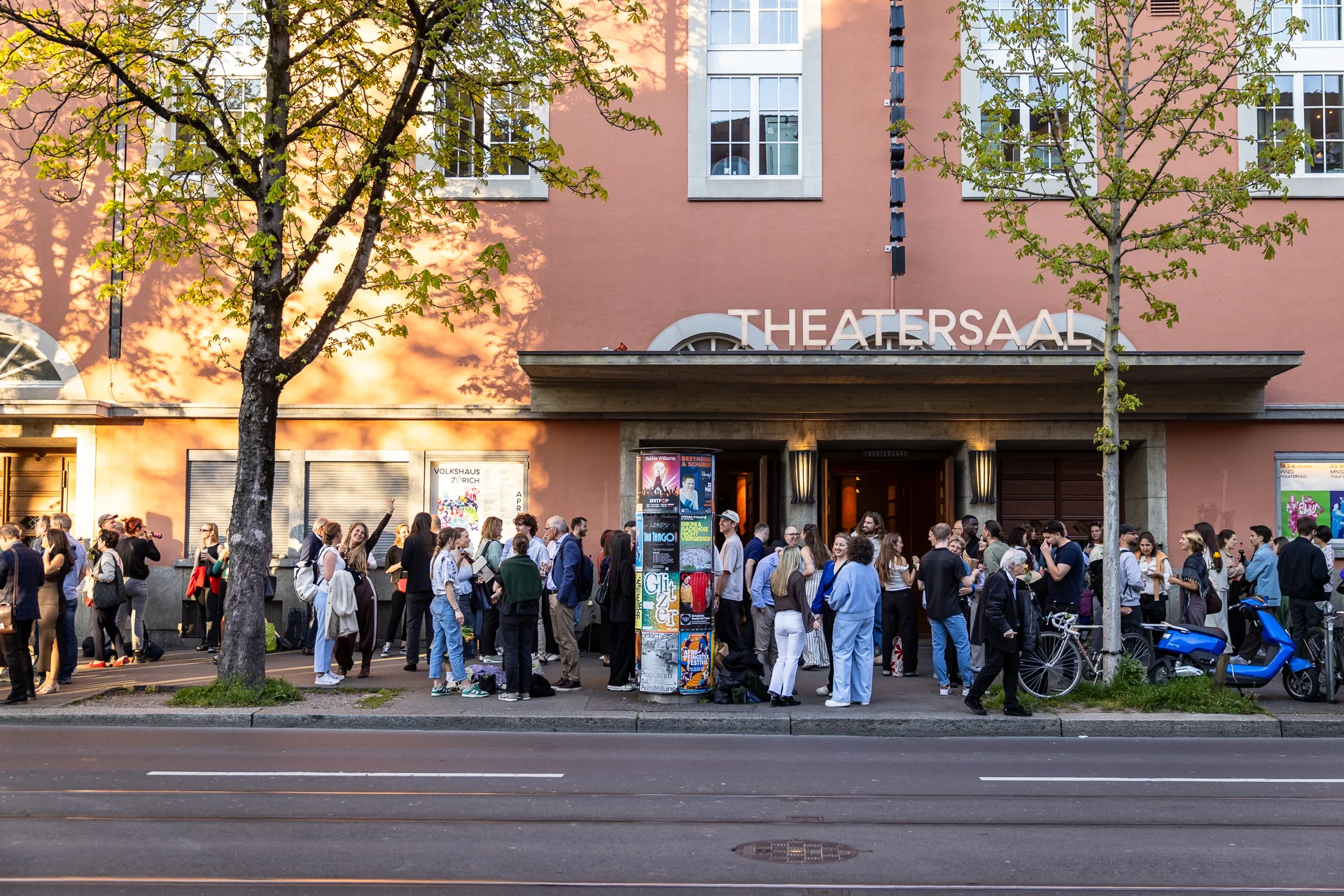 Blick von der anderen Strassenseite auf Absolvent:innen und Begleitpersonen. Sie stehen gut gelaunt vor dem Volkshaus in der Abendsonne.