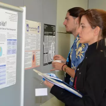 Konferenzbüro. Zwei Teilnehmende stehen vor einer Stellwand mit einem Poster der Poster-Session. Im Hintergrund weitere Poster, die an der Wand des Raums aufgehängt sind.