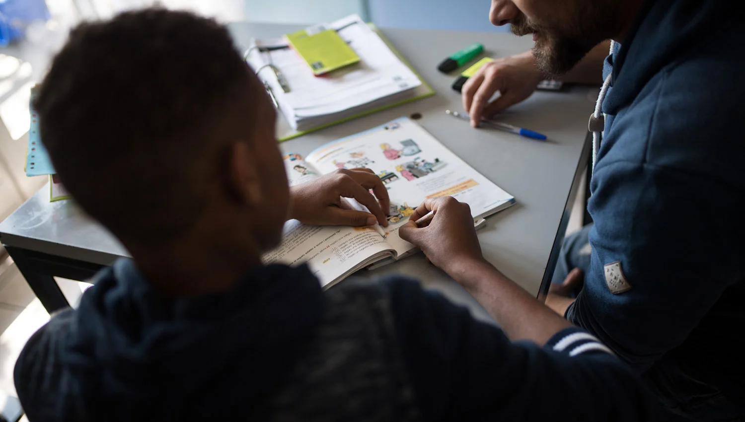 Ein Erwachsener unterstützt ein Kind am Tisch beim gemeinsamen Betrachten und Besprechen eines bebilderten Schulbuchs.