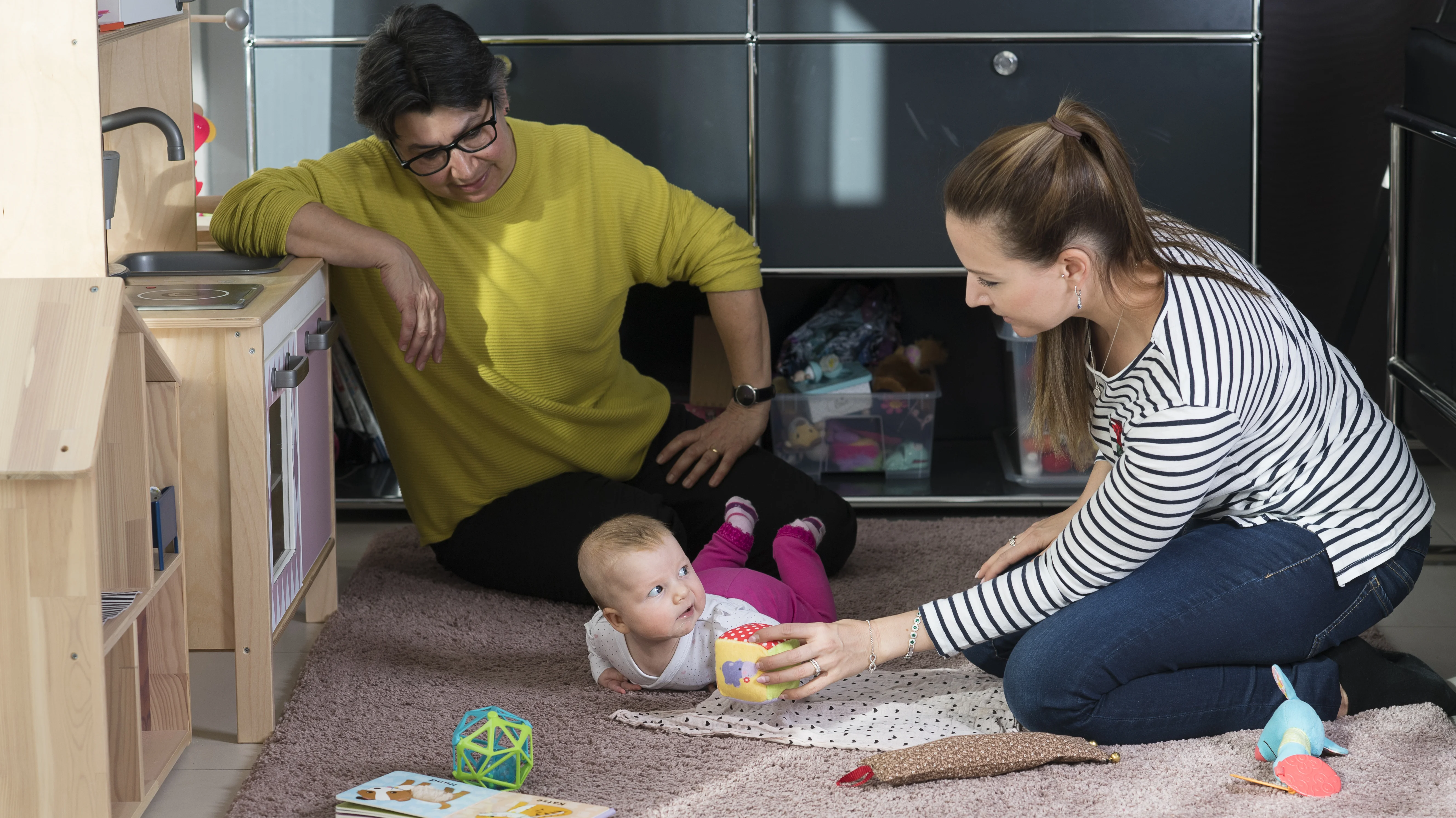Zwei Frauen die ein Kleinkind betreuen, auf dem Boden des Wohnzimmers sitzend.