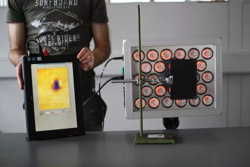 Das Bild zeigt die zu untersuchende Platte vor mehreren Halogenlampen und eine PErson die einen Computer hält auf den das Wärmebild angezeigt wird.