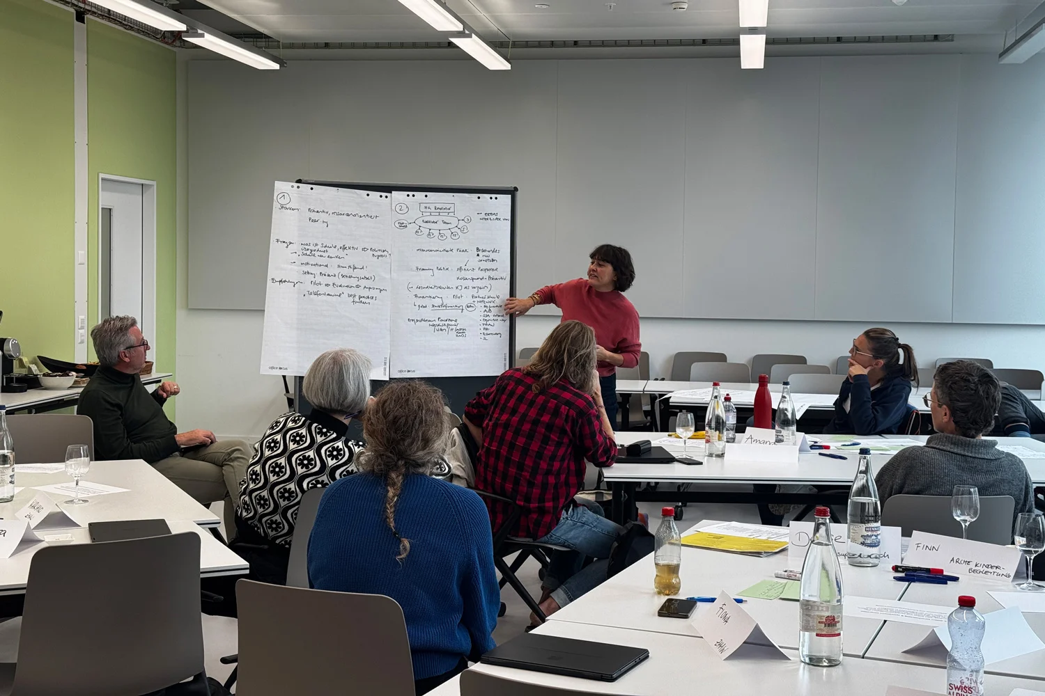 Im Unterrichtsraum sind Teilnehmende eines Workshops zu sehen, die an ihren Tischen sitzen. Vorne steht eine Frau mit rotem Pullover an einem Flipchart und stellt etwas vor.