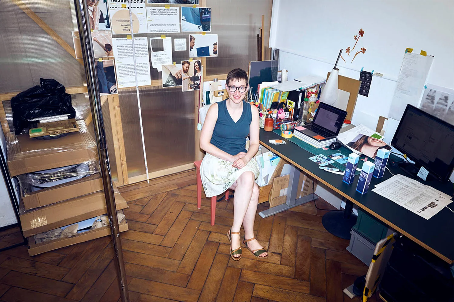 Sonja Koch sitzt lächelnd an ihrem Arbeitsplatz in einem Büro sitzt. Um sie herum befinden sich Computer, Unterlagen und Arbeitsmaterialien.