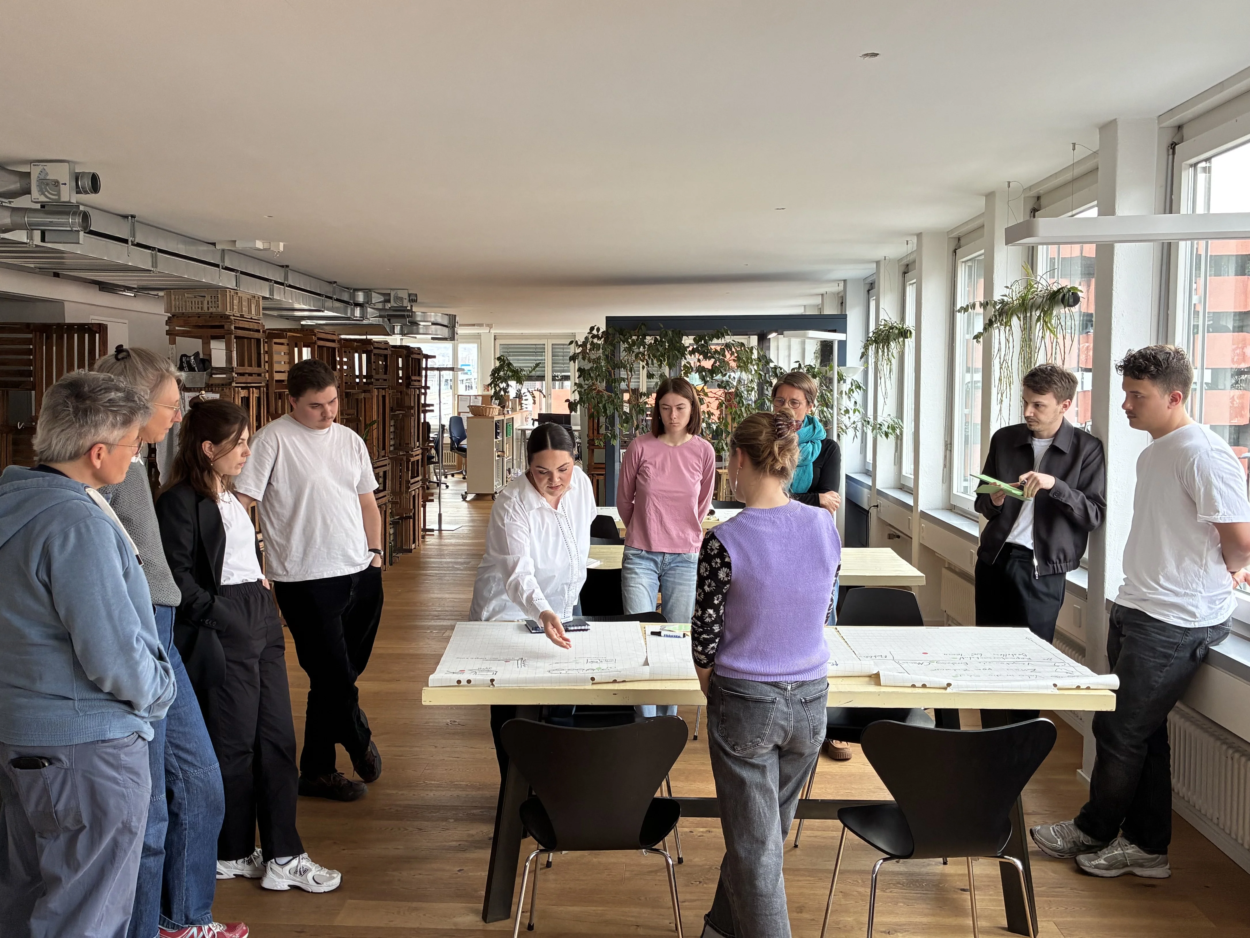 Eine Gruppe von Studierenden und Dozierenden steht um einen grossen Tisch in einem hellen, langgestreckten Raum mit grossen Fenstern. Auf dem Tisch liegen Papiere, im Hintergrund sind Pflanzen und Holzmöbel zu sehen