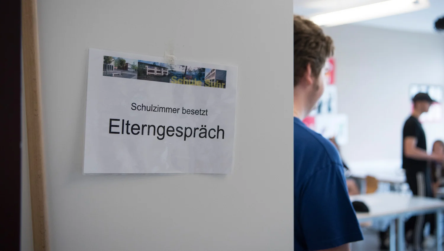 Leicht offene Schulzimmertüre. Im Zimmer sieht man einen Schüler und dessen Lehrer oder ein Elternteil.