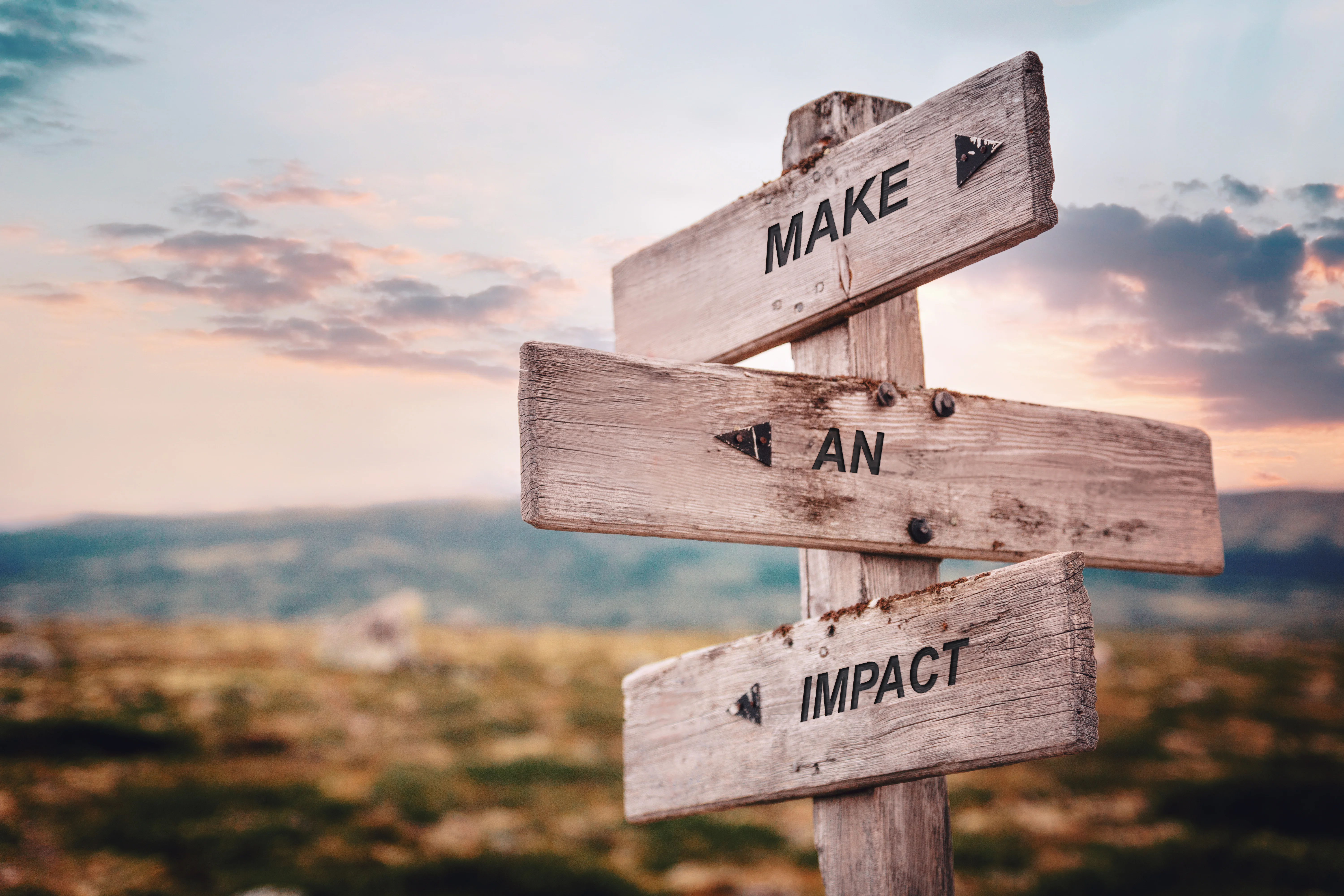 Wooden signpost in an open landscape. The three signs display the words “MAKE”, “AN”, and “IMPACT”, symbolizing guidance, impact, and positive change.