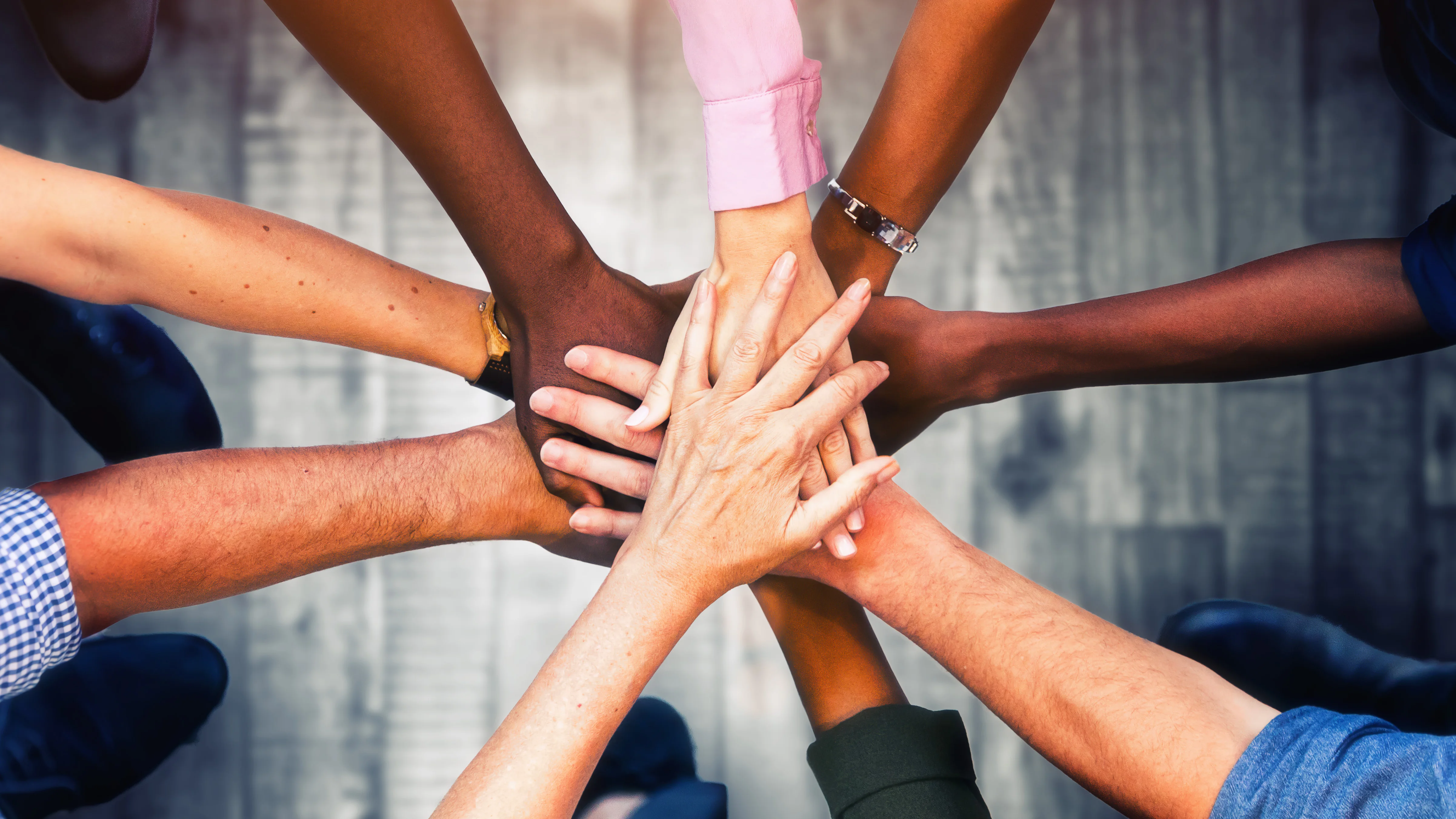 Several people of different skin tones stack their hands together in the center of the frame, photographed from above.