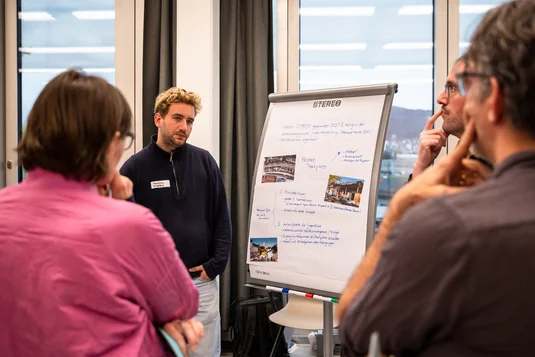 Bild vergrössern: Eine Person stellt zwei Workshop-Teilnehmenden an einem Flipchart ein Projekt aus dem Sozialbereich vor
