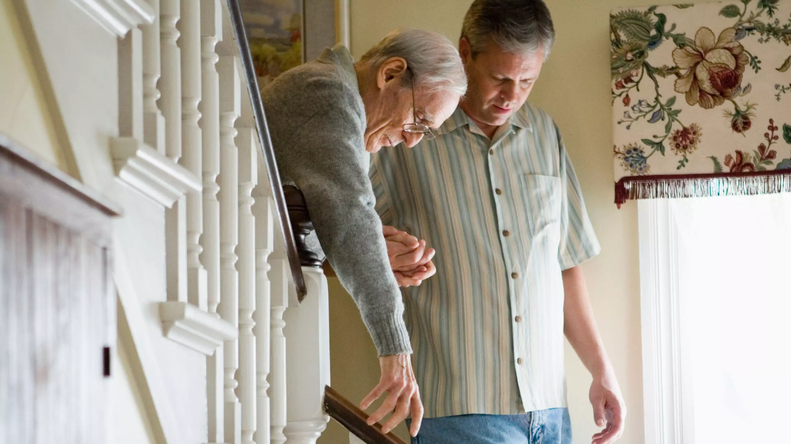 Alter Mann wird gestützt von einem etwas jüngeren Mann beim Hinuntergehen einer Treppe im Haus