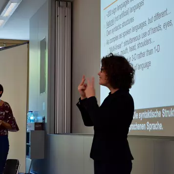 Hörsaal. Irene Strasly trägt ihre Präsentation vor, eine Gebärdendolmetscherin übersetzt, beide Frauen stehen.