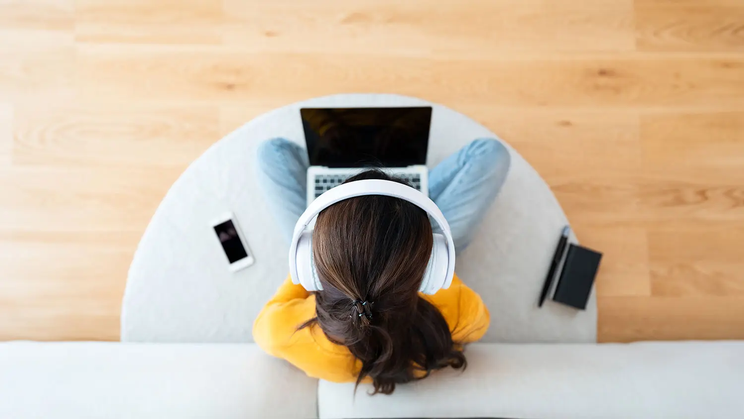 Top view of young Asian woman sitting on the floor and working on computer laptop with book, pen and mobile smartphone, wearing wireless headset, listening to music