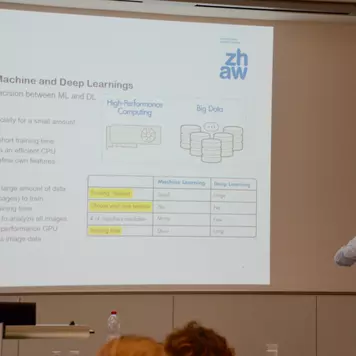 Hörsaal. Andri Reichenbacher präsentiert seinen Vortrag. Im Hintergrund eine seiner Vortragsfolien. Sie trägt den Titel: Practicality. Comparision of Machine and Deep Learnings.
