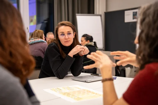 Bild vergrössern: Eine Teilnehmende eines Workshops sitz am Gruppentisch und hört einer anderen Person zu