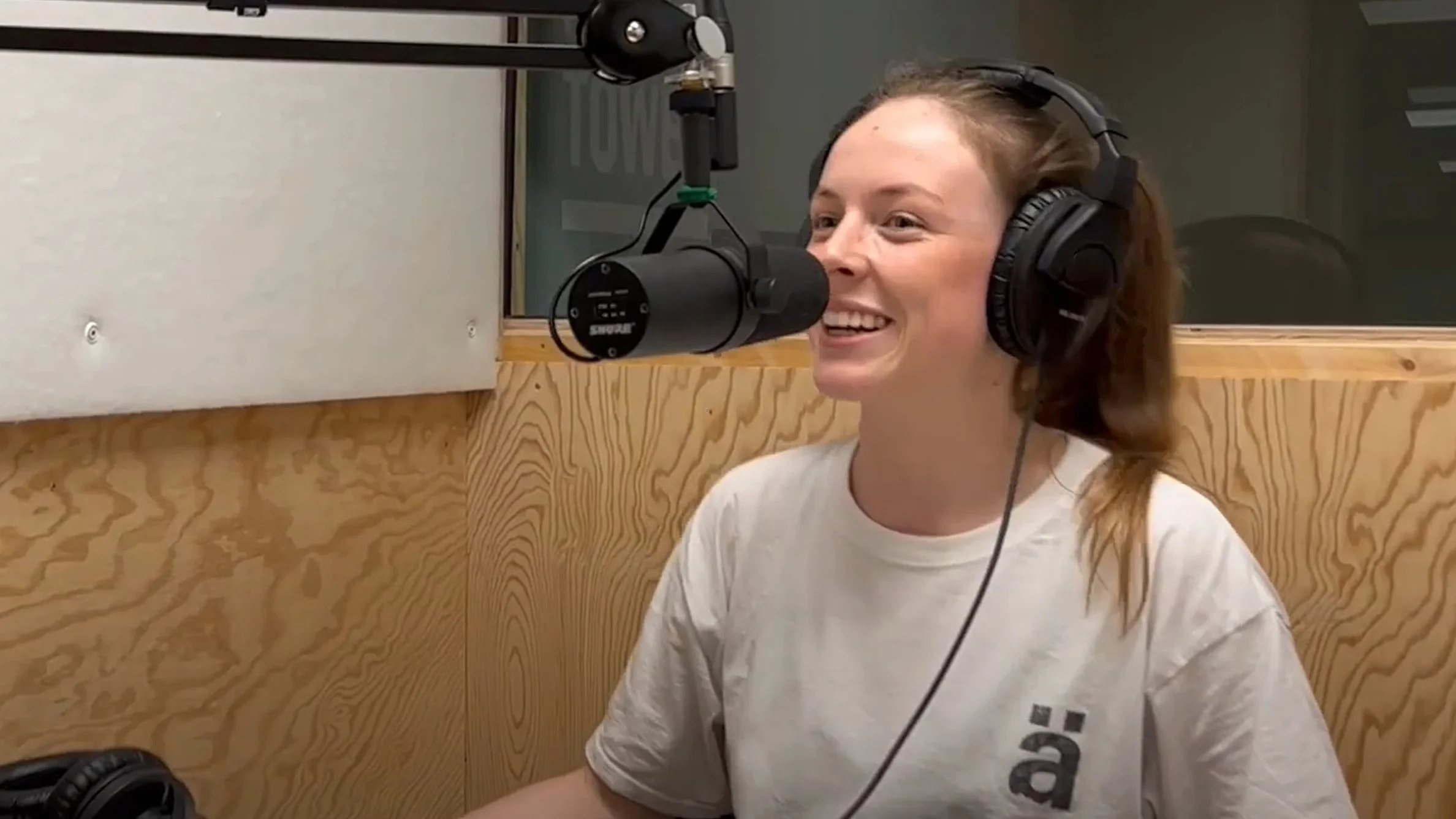 Lächelnde Person mit Kopfhörern spricht in ein Mikrofon in einem Tonstudio mit Holzverkleidung.