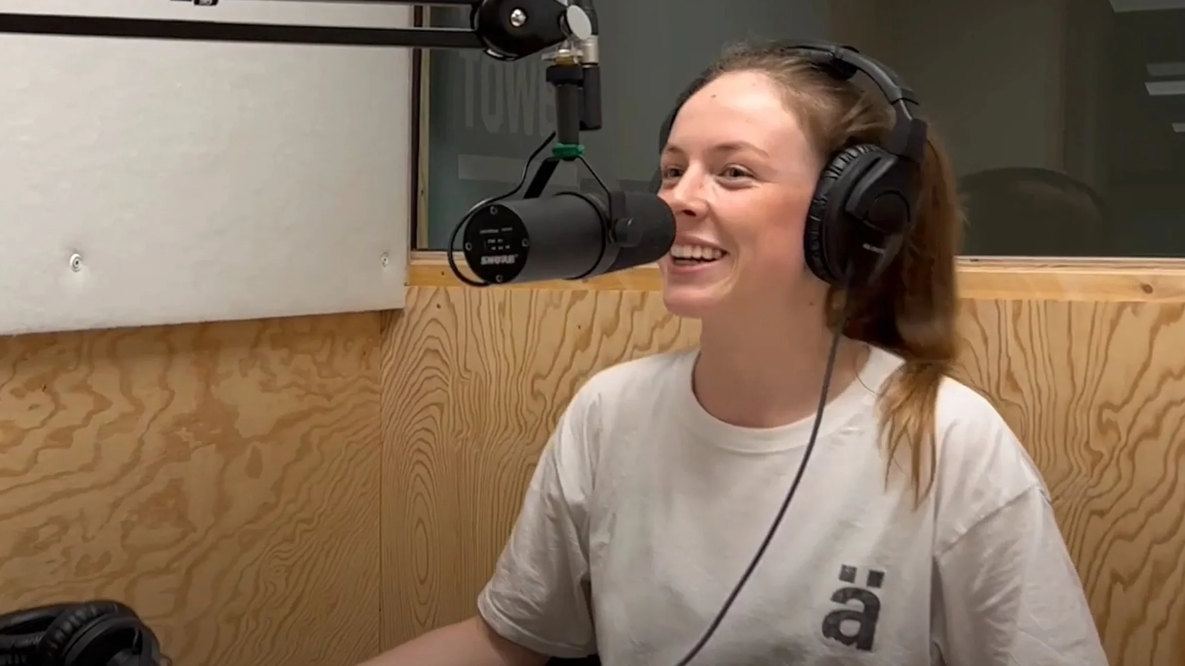 Smiling person wearing headphones speaks into a microphone in a wood-paneled recording studio.