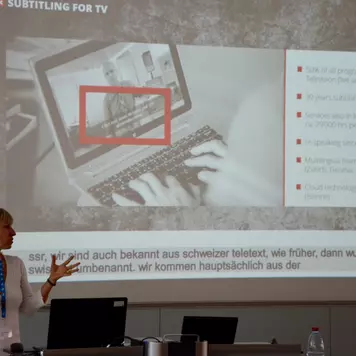 Hörsaal. Michaela Nachtrab präsentiert ihren Vortrag. Im Hintergrund eine ihrer Vortragsfolien mit dem Titel: Subtitling for TV.