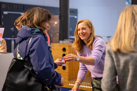 Bild vergrössern: Mitarbeiterin am Empfang begrüsst teilnehmende Person und händigt Namensschild aus