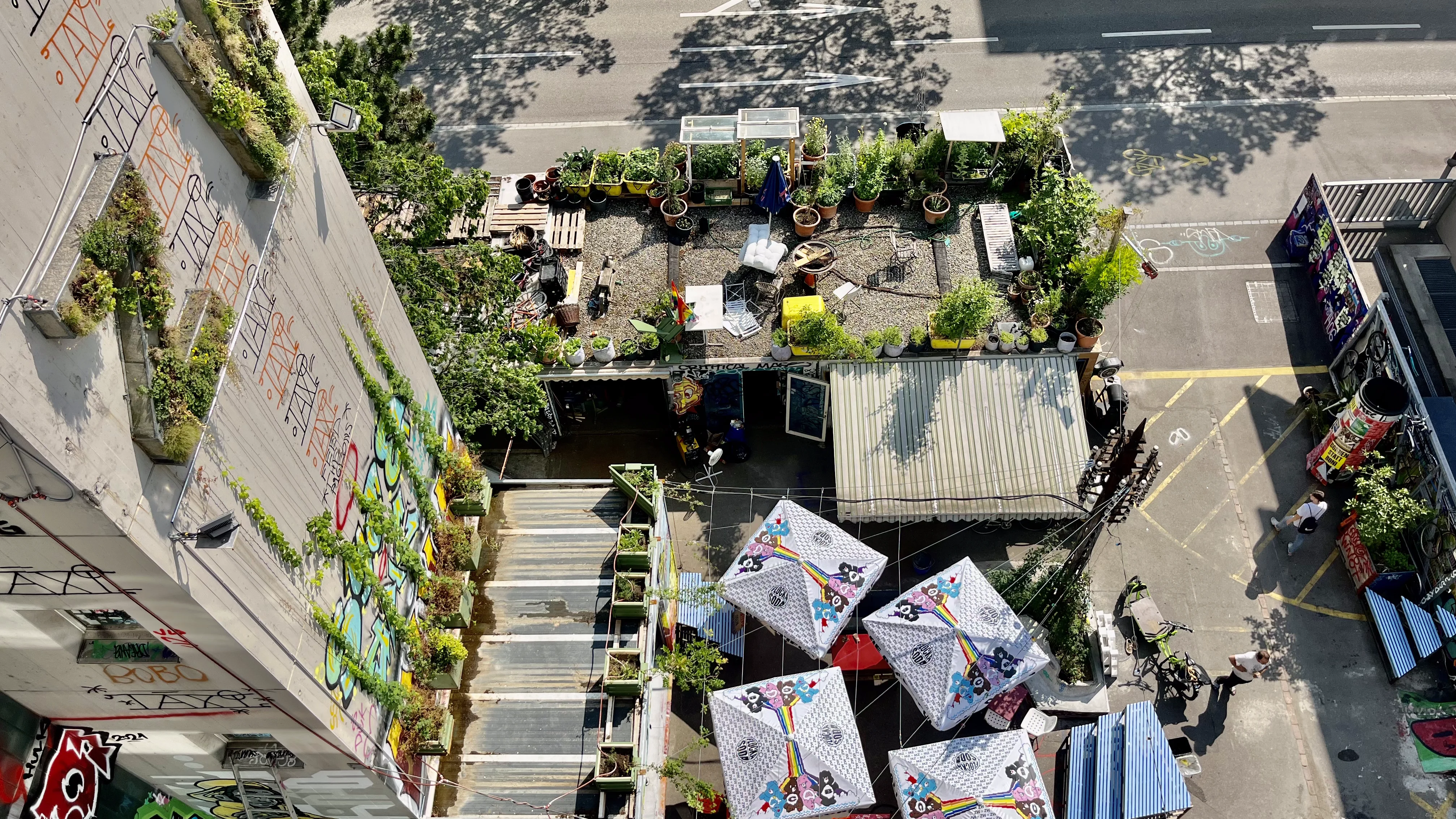 Vogelperspektive auf einen begrünten Innenhof zwischen Gebäuden mit Pflanzen, Sonnenschirmen und einer bemalten, graffitiverzierten Fassade an einer Strasse.