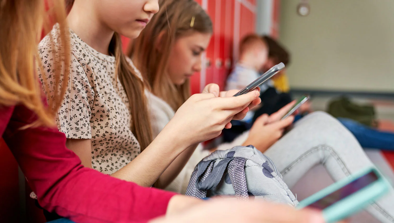 Mehrere Kinder sitzen in einem Schulflur vor Schließfächern und schauen auf ihre Smartphones.