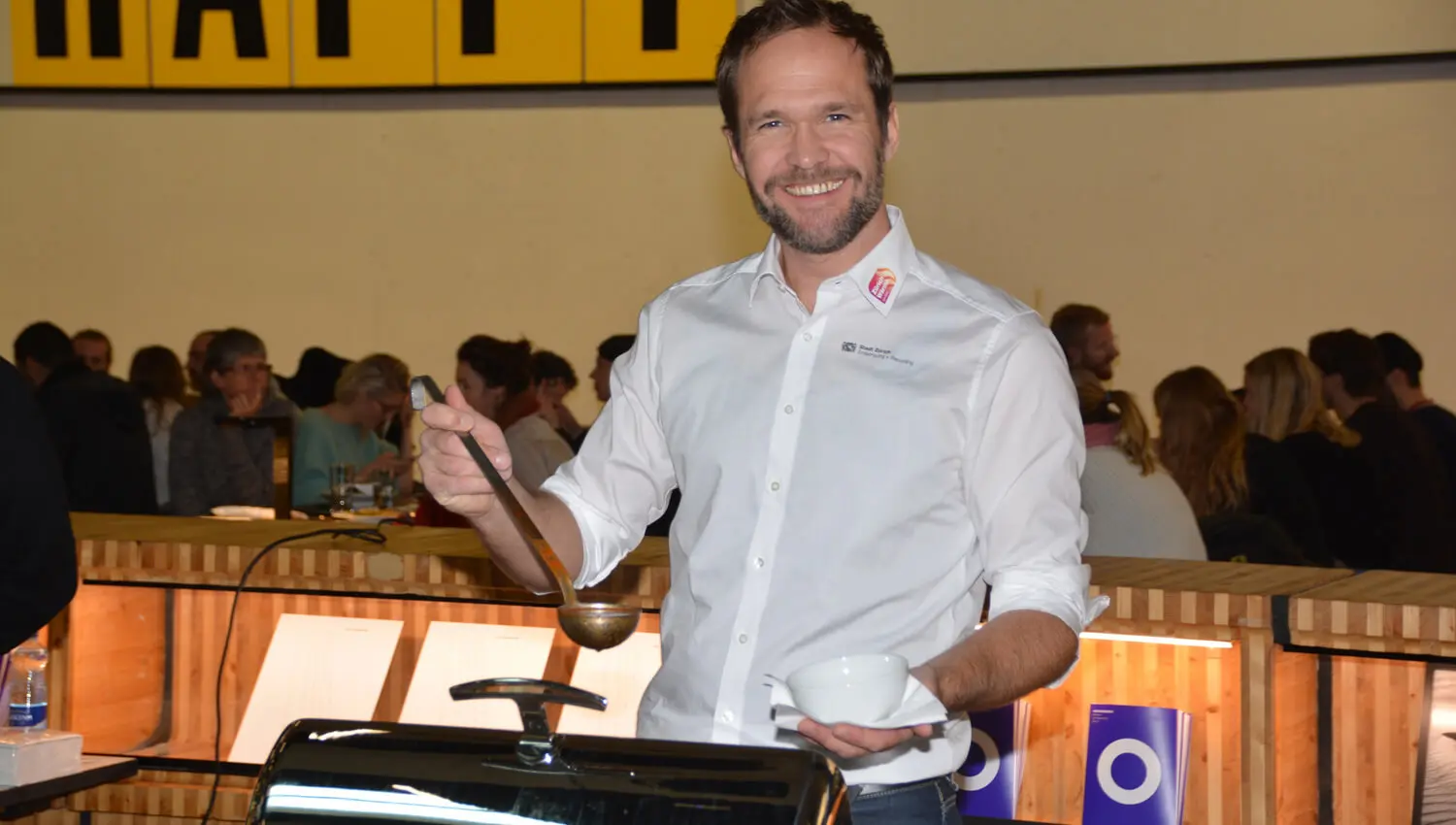 Ein lächelnder Herr mit einer Suppenkelle steht in der Eingangshalle des Toni-Areals und schöpft Suppe.
