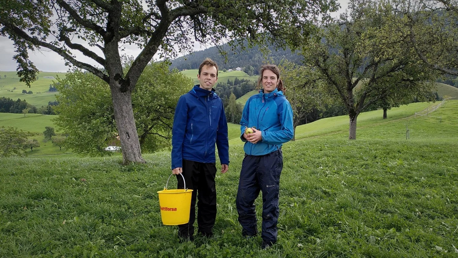 Zwei Personen in Outdoor-Jacken stehen auf einer grünen Hügellandschaft unter einem Apfelbaum, eine hält einen gelben Eimer, im Hintergrund weite Wiesen und Hügel