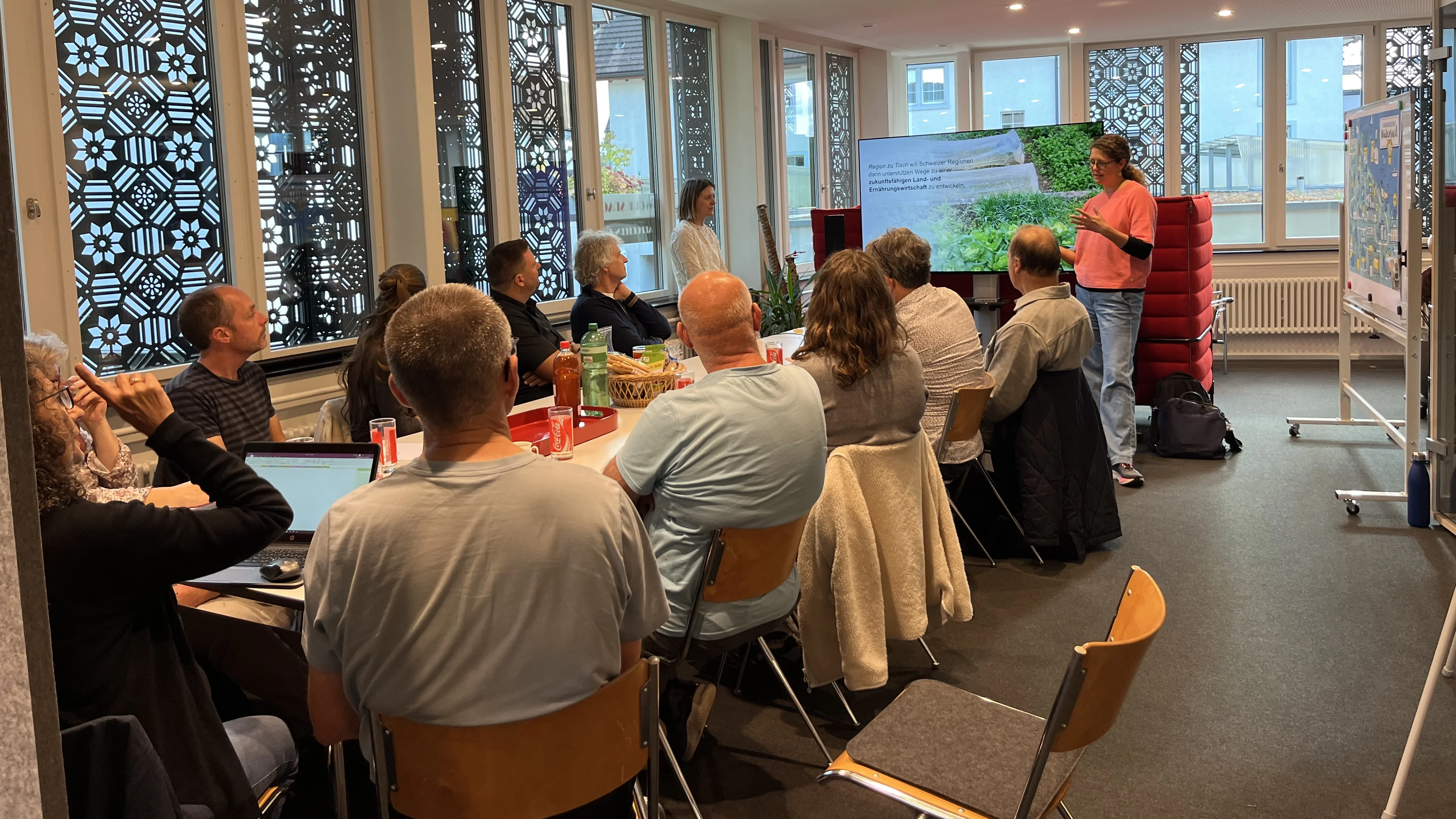 Auf dem Bild sieht man die die Gruppe vom ersten Infoabend um einen runden Tisch sitzen und der Projektvorstellung zuhören.