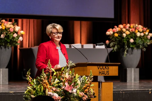 Bild vergrössern: Die stellvertretende Direktorin des Departements, Monika Götzö, hält auf der Bühne die Begrüssungsrede. Die Bühne ist mit Blumen dekoriert.