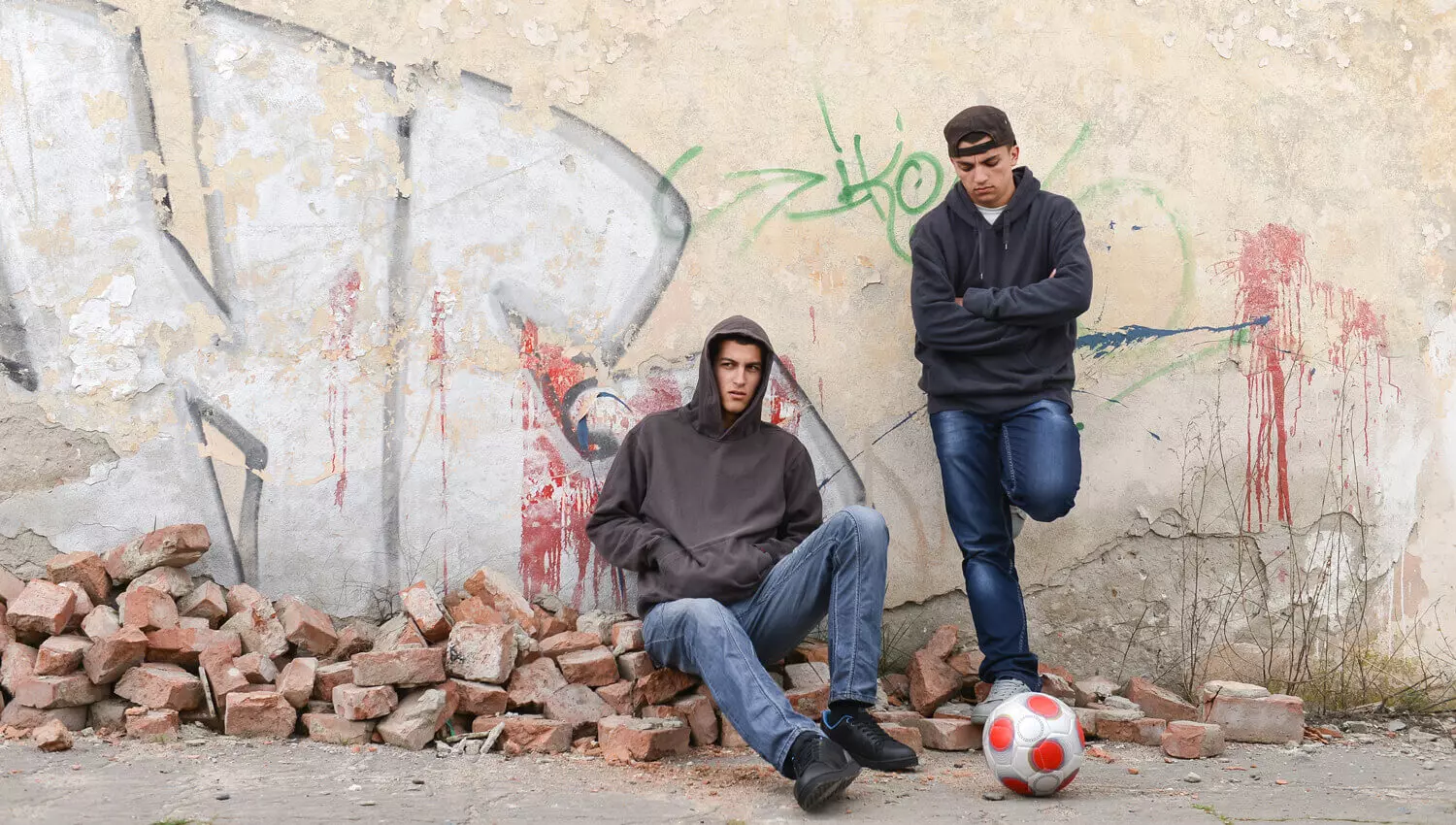 Ein Jugendlicher lehnt an eine Wand, der eine sitzt daneben auf dem Boden mit dem Rücken zur Wand. 