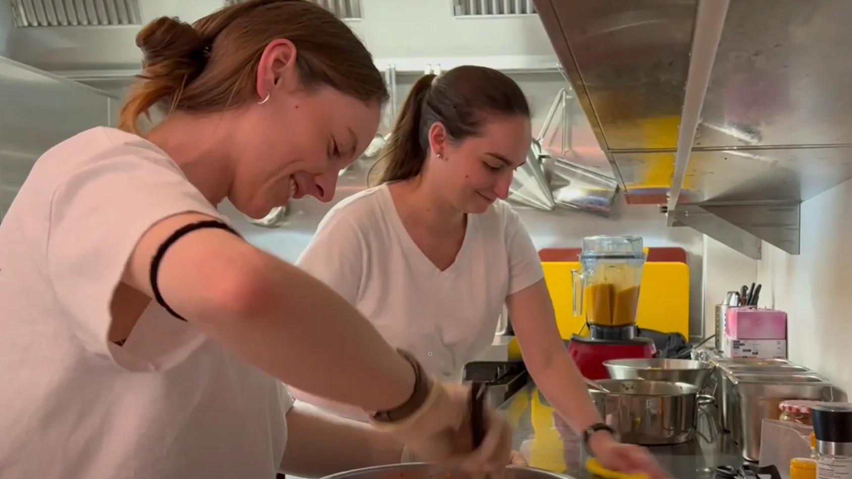 Zwei Personen in einer Küche bereiten lächelnd Speisen zu, im Hintergrund ein Mixer und Kochtöpfe.