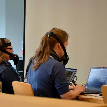 Hörsaal. Rückansicht der beiden Schriftdolmetscherinnen während der Arbeit. Sie tragen Stenomasken und haben ihre Laptops vor sich auf dem Tisch.