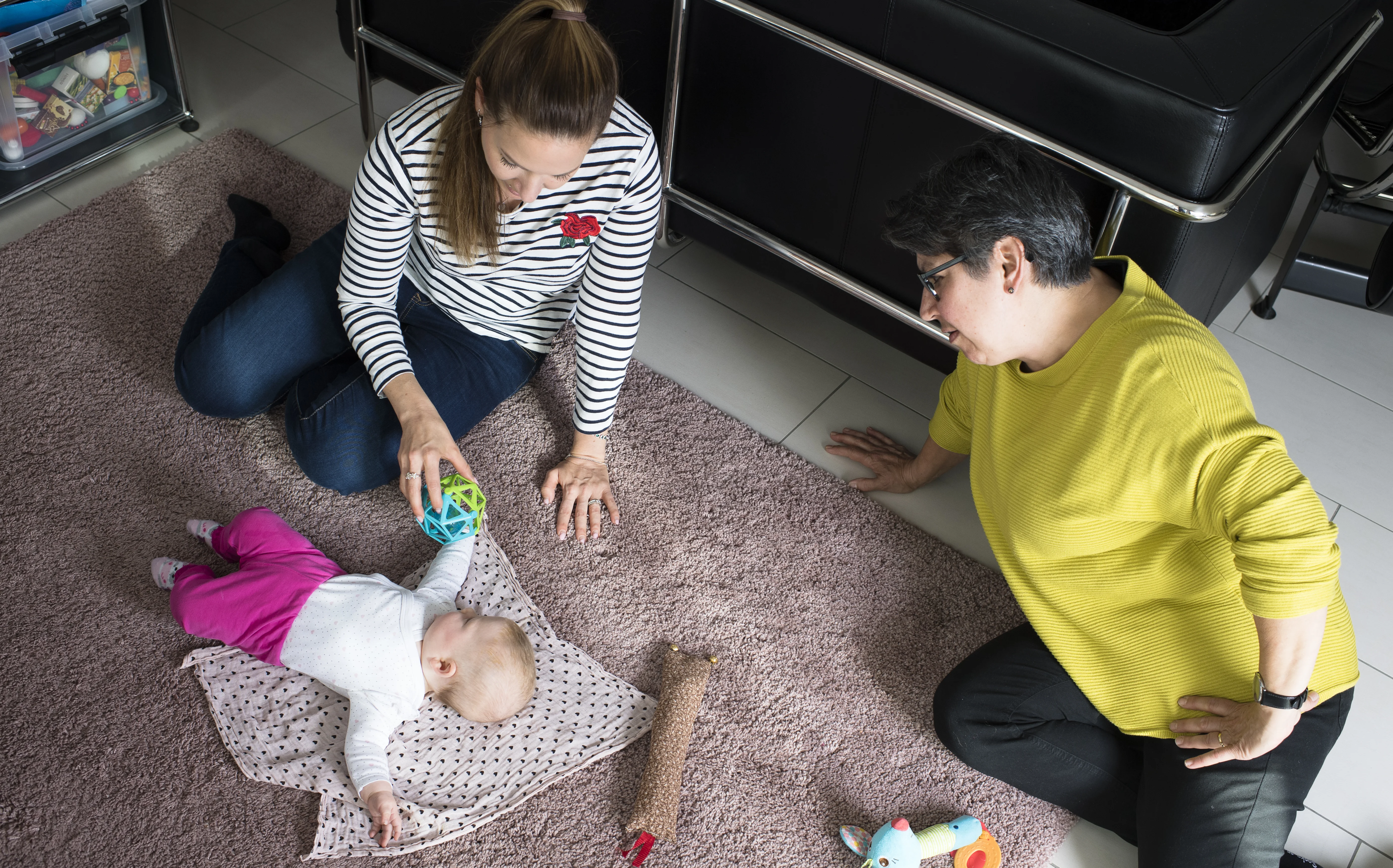 Zwei Frauen die ein Kleinkind betreuen, auf dem Boden des Wohnzimmers sitzend.
