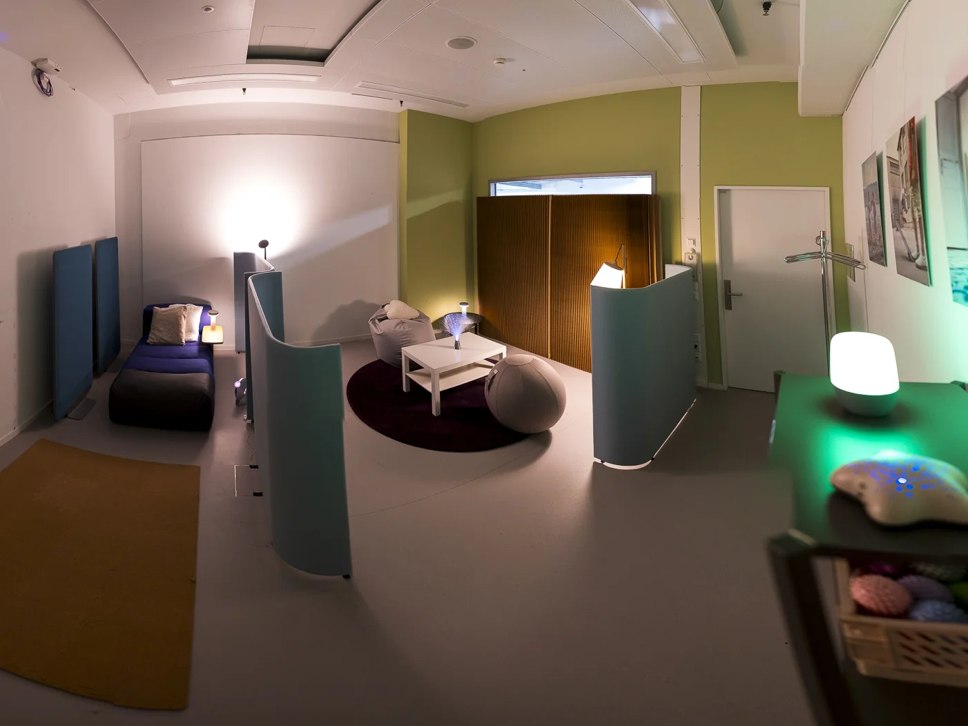 Panoramaaufnahme eines Sensory Rooms mit gedämpftem, warmem Licht. Ein niedriges Bett mit dunkelblauer Decke und ein kleiner weisser Tisch mit Bodenkissen stehen im Raum, getrennt durch geschwungene Trennwände. Rechts leuchtet eine Lampe in Grün und Weiss, in der Mitte liegt ein Sitzsack.