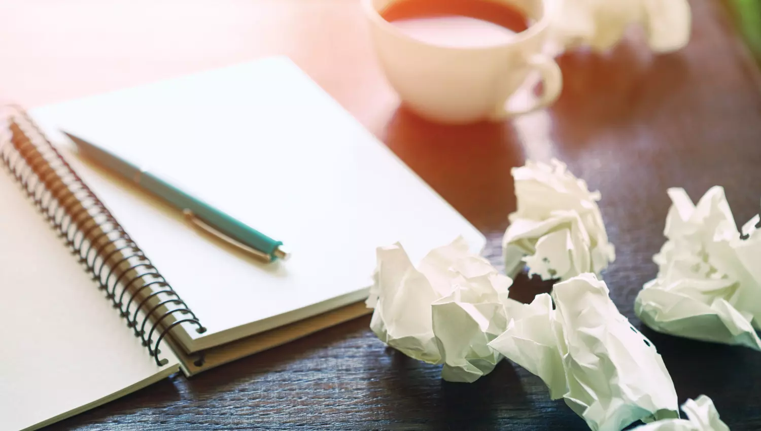 Ein aufgeschlagenes Notizbuch, zerknüllte Papierballen und eine Tasse Kaffee stehen auf einem Tisch.