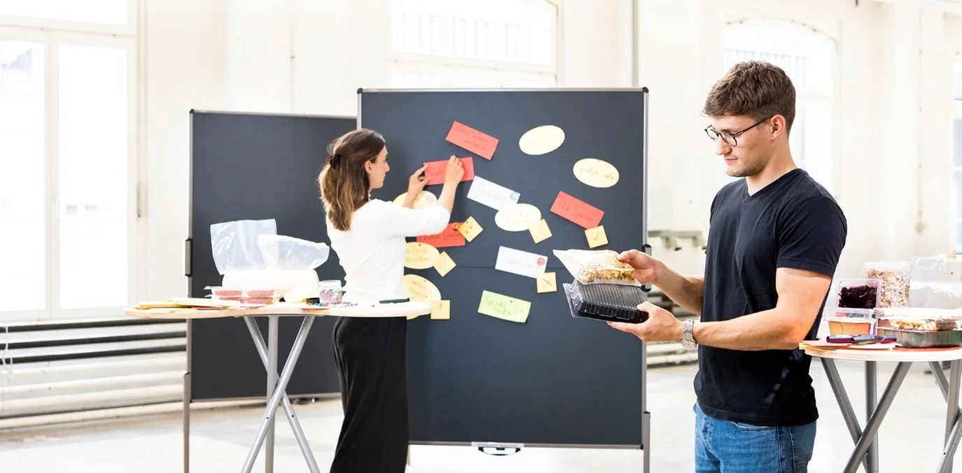 Two people are working in an innovation workshop with food items and sticky notes on a pinboard.