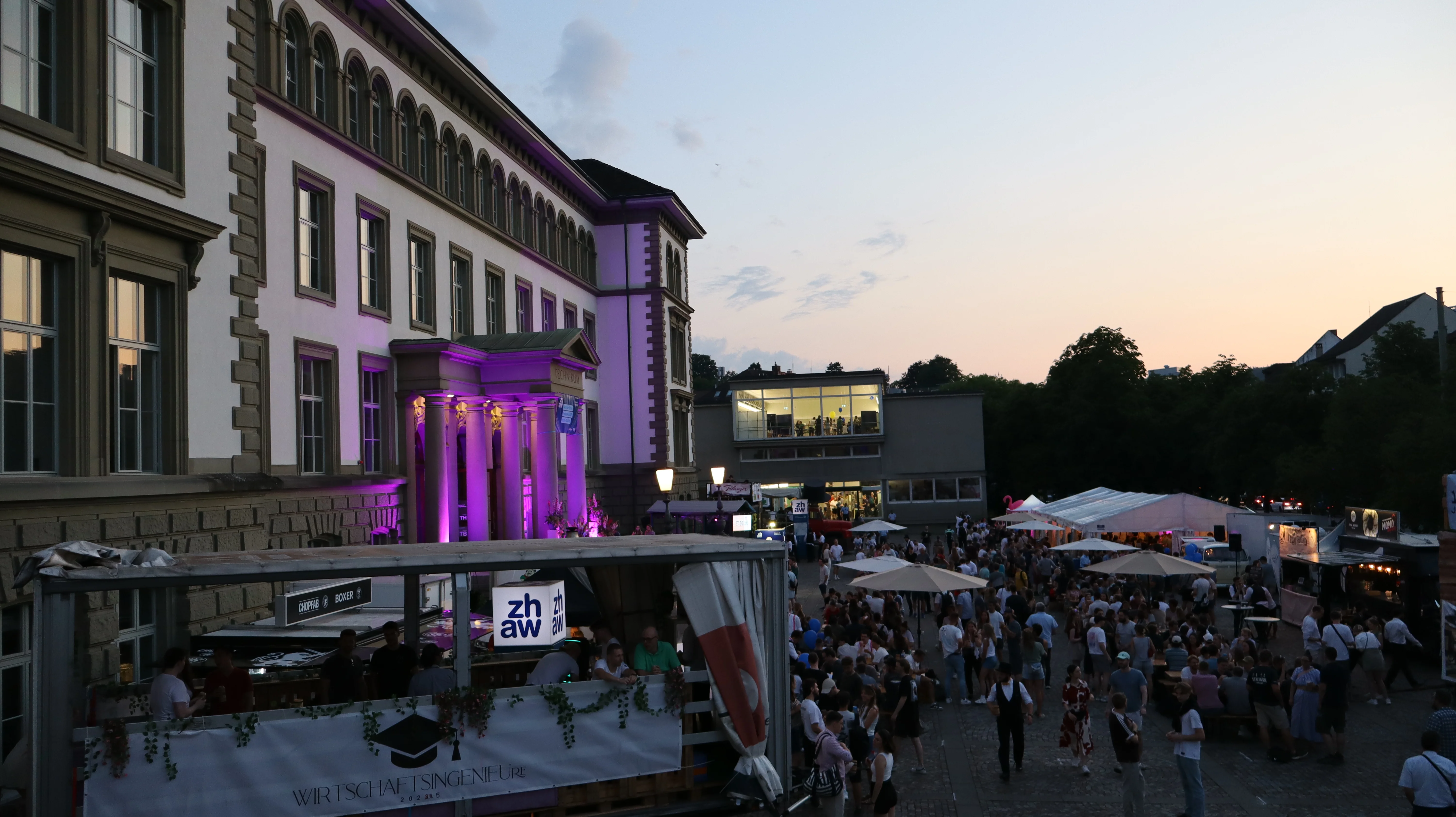Blick auf den Vorplatz bei untergehenden Sonne. Zu sehen sind viele Leute ein grosses Zelt und Sonnenschirme.
