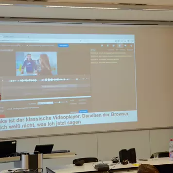 Hörsaal. Christian David und Alexander Kurch präsentieren ihren Vortrag. Im Hintergrund eine ihrer Vortragsfolien auf der ein Screenshot der Software, welche die beiden vorstellen, sichtbar ist.