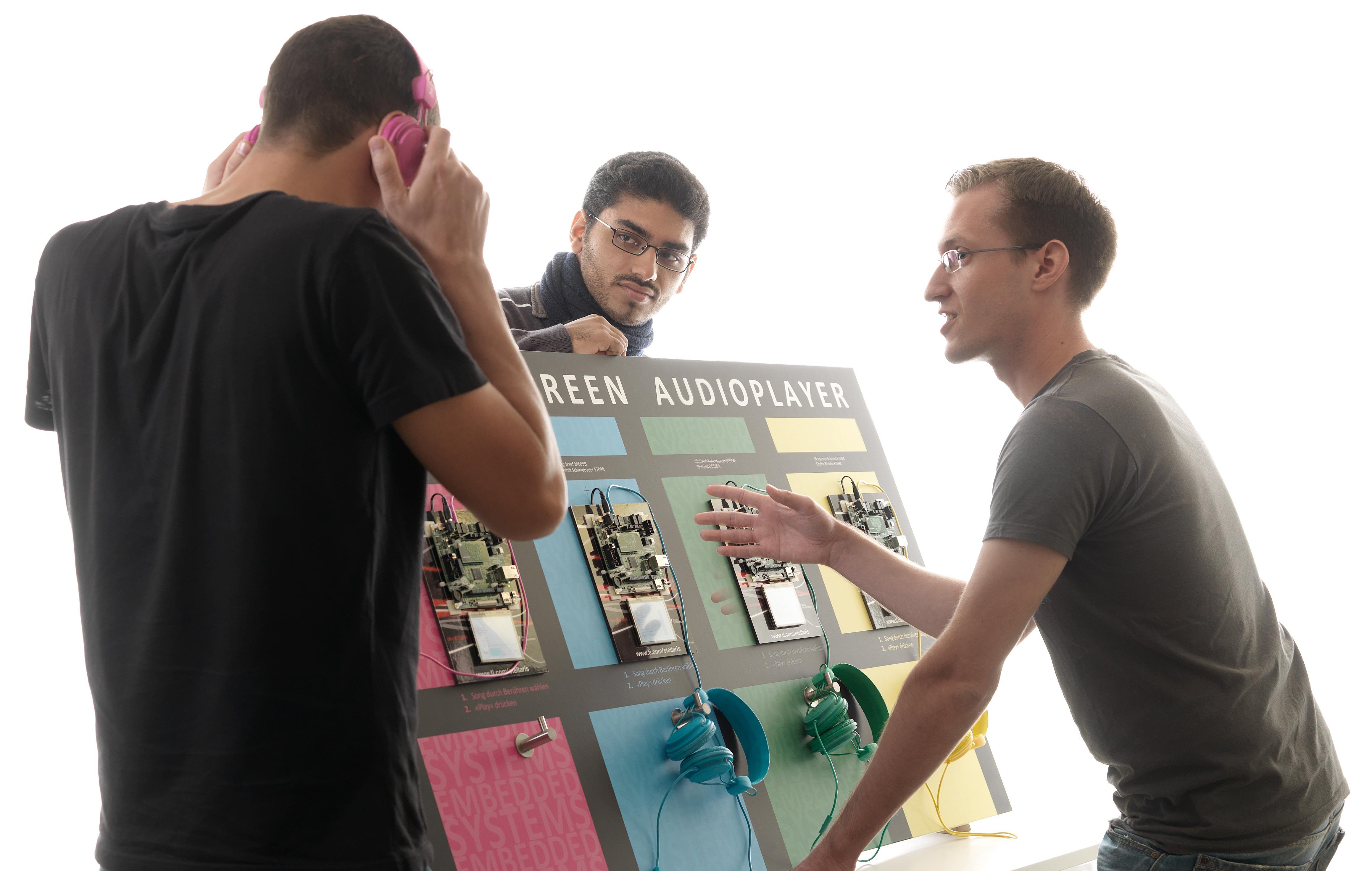 Das Bild zeigt drei Studierende. Einer erklärt einem anderen eine FPGA-basierte Versuchsanlage für Tonübertragung und ein dritter schaut dabei zu.