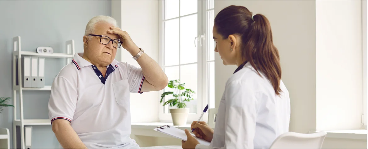 Senior man patient suffering from a headache, sitting on a medical couch, holding hand on his head and telling the doctor about the intense pain that he is feeling