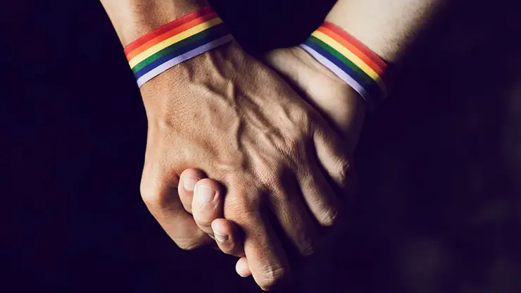 Zwei Hände halten sich. Am Handgelenk tragen beide Hände eine Regenbogen-Armband.