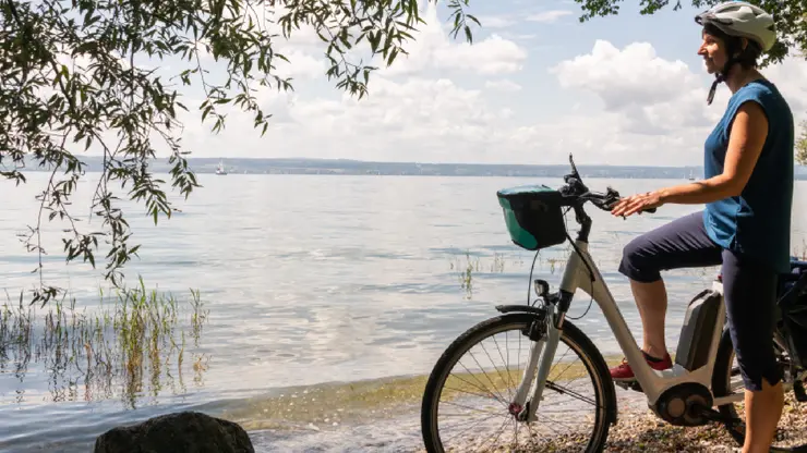 Eine Frau steht beim Fahrrad am Seeufer und schaut auf den See.