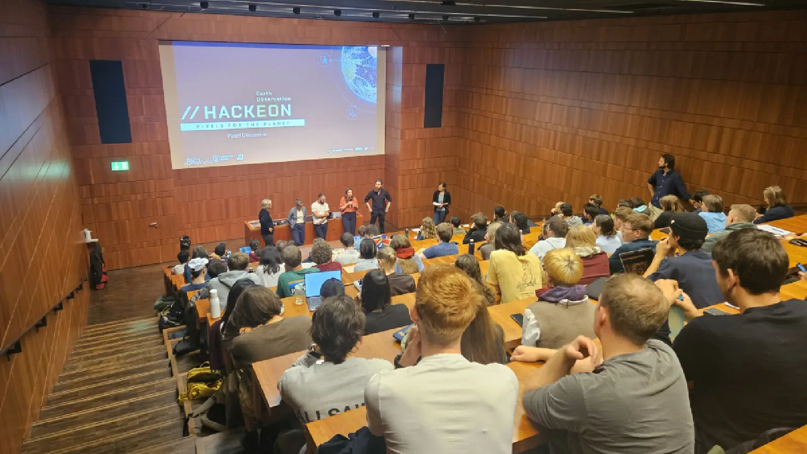 Studierende im Hörsaal hören einer Diskussion von fünf Personen zu