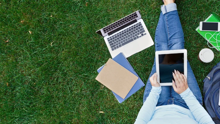 Sicht von oben: Eine Frau sitzt mit ausgestreckten Beinen auf einer Wiese. Sie arbeitet am Tablet, ein Note ist auf der Wiese, ebenso ein Rucksack, die Sonnebrille, ein Notizbuch und das Handy.